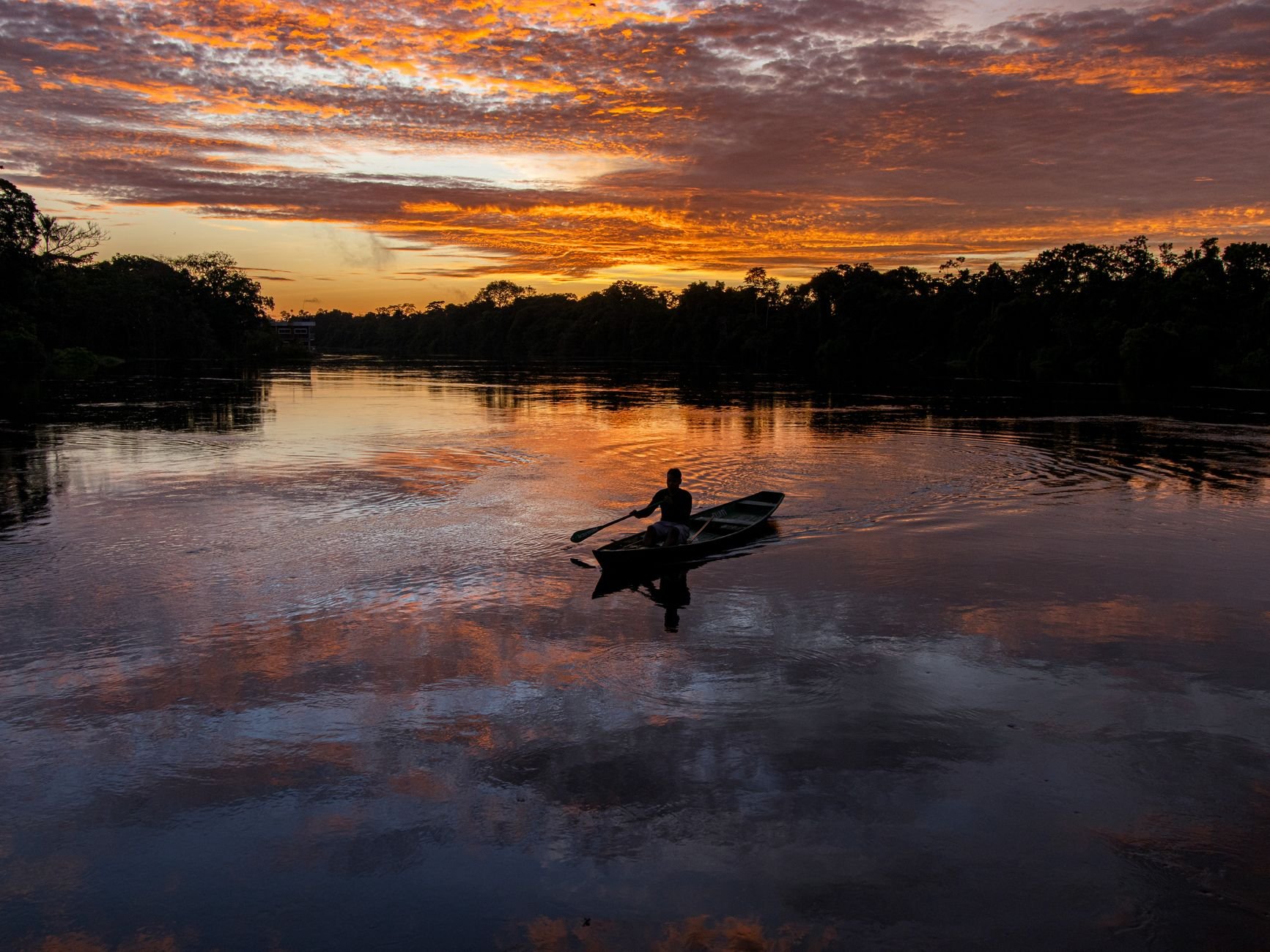 A fisherman rows a boat across the Amazon River at sunset, surrounded by vivid reflections